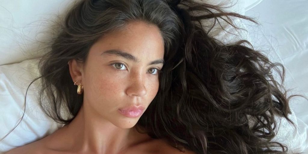Woman with dark, wavy hair lying on a bed, looking up at the camera.