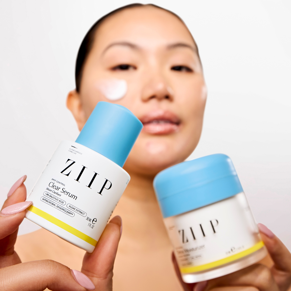 Woman holding ZIIP Clear serum and Detox moisturizer bundle with product applied to cheek
